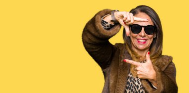 Güzel orta yaş zarif kadın gözlük ve vizon ceket giyen mutlu yüz ile eller ve parmaklar ile çerçeve yapma gülümseyen. Yaratıcılık ve Fotoğrafçılık konsepti.