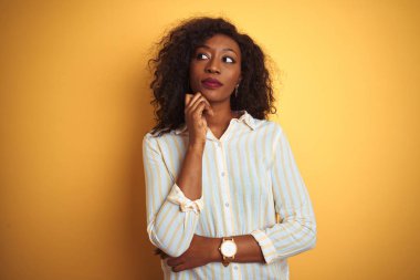 Çizgili gömlek giyen Afro-amerikan kadın izole sarı arka plan üzerinde duran çene üzerinde soru, dalgın ifade düşünme. Düşünceli bir yüzle gülümsüyor. Şüphe kavramı.