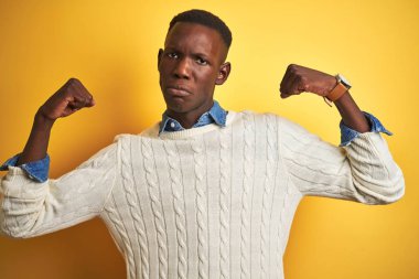 Kot gömlek giyen Afrikalı Amerikan adam ve izole sarı arka plan üzerinde beyaz kazak gurur gülümseyen silah kasları gösteren. Fitness konsepti.