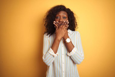 Izole sarı arka plan üzerinde duran çizgili gömlek giyen Afro-Amerikan kadın hata için elleri ile ağız kapsayan şok. Gizli kavram.