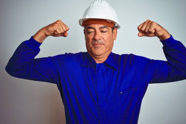 Yakışıklı orta yaş Worker adam üniforma ve kask üzerinde izole beyaz arka plan gösteren silah kasları gururlu gülümsüyor giyiyor. Fitness konsepti.
