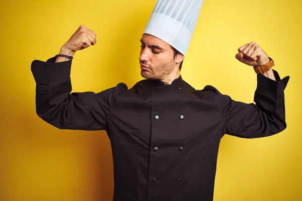 Young chef man wearing uniform and hat standing over isolated yellow background showing arms muscles smiling proud. Fitness concept.