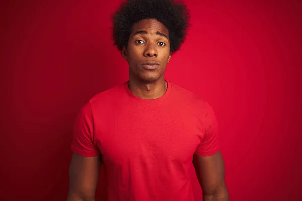 Young american man with afro hair wearing t-shirt standing over isolated red background with serious expression on face. Simple and natural looking at the camera.