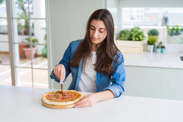 Beautiful young woman cutting a tasty pizza slice using a cutter with a confident expression on smart face thinking serious