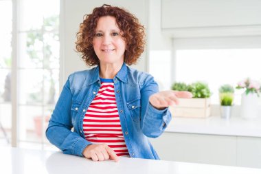 Evde kot ceket giyen kıvırcık saçlı orta yaş yaşlı kadın yardım ve kabul veren neşeli sunan palmiye el gülümseyerek.