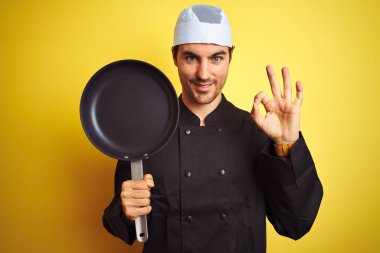Üniforma giyen ve şapka tutan genç şef adam izole sarı arka plan üzerinde parmaklarla ok işareti yapıyor, mükemmel sembolü