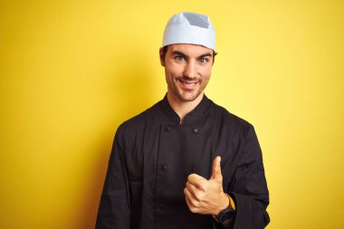 Genç yakışıklı Şef adam pişirerek üniforma ve şapka üzerinde izole sarı arka plan yapıyor mutlu thumbs yukarı jest ile el. Başarı göstererek kameraya bakarak ifadeyi onaylama.