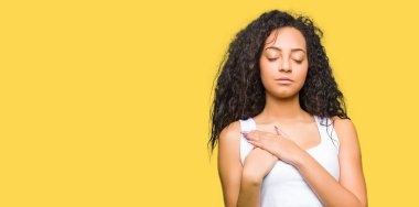 Kıvırcık saçları ile gülümseyen genç güzel kız göğüs kapalı gözler ve yüzünde minnettar jest ile eller. Sağlık kavramı.