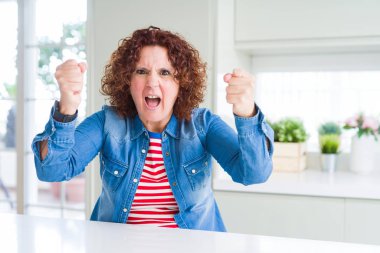 Evde kot ceket giyen kıvırcık saçlı orta yaş yaşlı kadın kızgın ve öfkeli öfke ile bağırırken sinirli ve öfkeli yumruklar yükselterek. Öfke ve agresif kavram.