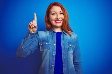 Mavi izole arka plan üzerinde ayakta kot gömlek giyen genç güzel Redhead kadın gösteren ve emin ve mutlu gülümseyen iken parmak numarası bir ile işaret.