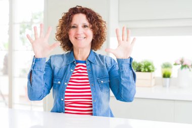 Kıvırcık saçlı orta yaş yaşlı kadın evde kot ceket giyen gösteren ve parmakları ile işaret ederken emin ve mutlu gülümseyerek on numara.