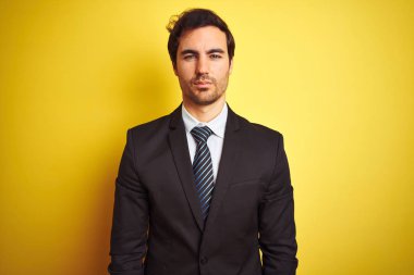 Genç yakışıklı işadamı giyen takım elbise ve kravat ayakta izole sarı arka plan yüzünde ciddi bir ifade ile rahat. Basit ve doğal kamera bakıyor.
