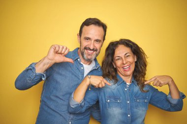 Güzel orta yaş çift birlikte izole sarı arka plan üzerinde ayakta yüz gülümseme ile kendine güvenen görünümlü, parmaklarıyla gurur ve mutlu kendini işaret.