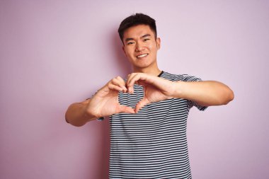 Genç Asyalı Çinli adam izole pembe arka plan üzerinde ayakta çizgili t-shirt giyerek kalp sembolü ve elleri ile şekil gösteren aşk gülümsüyor. Romantik konsept.