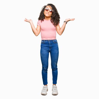 Kıvırcık saçlı pembe güneş gözlüğü clueless ve şaşkın bir ifadeyle kollar ve eller kaldırdı giyen genç güzel kadın. Şüphe kavramı.