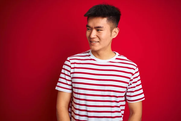孤立した赤い背景の上に立つストライプのTシャツを着たアジアの若い中国人男性は 笑顔で横に向かって 自然な表情 自信を持って笑う — ストック写真