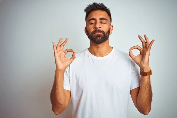 Young indian man wearing t-shirt standing over isolated white background relaxed and smiling with eyes closed doing meditation gesture with fingers. Yoga concept.