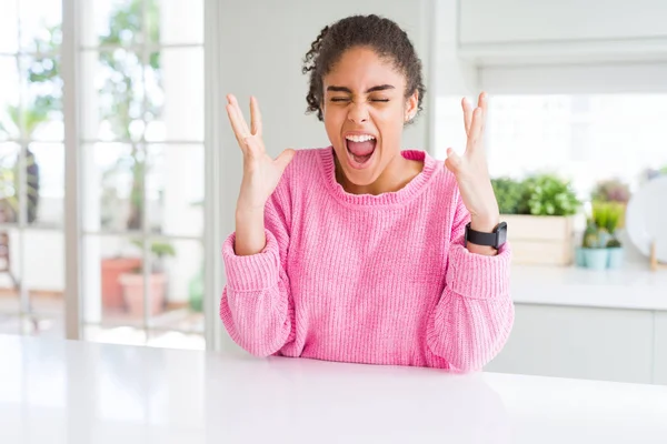 Beautiful african american woman with afro hair wearing casual pink sweater celebrating mad and crazy for success with arms raised and closed eyes screaming excited. Winner concept