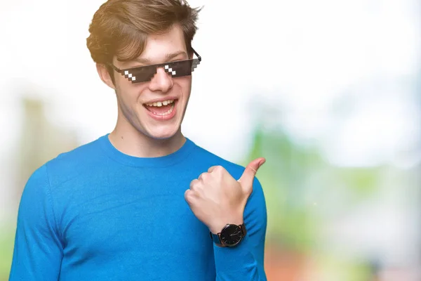 Young man wearing funny thug life glasses over isolated background smiling with happy face looking and pointing to the side with thumb up.