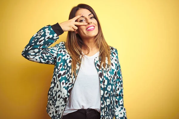 Sarı izole arka plan üzerinde rahat ceket giyen genç güzel kadın yüz üzerinde parmaklarla barış sembolü yapıyor, neşeli gösteren zafer gülümseyen