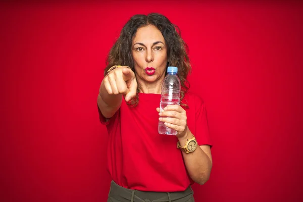 中年老妇人拿着塑料水瓶在红色孤立的背景用手指指着相机和你 手符号 积极和自信的姿态从前面 — 图库照片