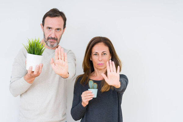 Пара средних лет держит растения на изолированном фоне с открытой рукой делает знак стоп с серьезным и уверенным выражением, защитный жест

