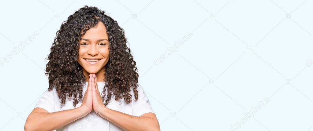 Young beautiful woman with curly hair wearing white t-shirt praying with hands together asking for forgiveness smiling confident.
