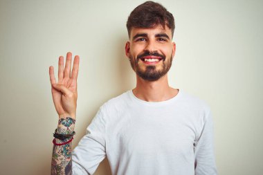 Dövme ile genç adam izole beyaz arka plan üzerinde ayakta t-shirt dikiliyor gösteren ve parmak sayısı dört ile işaret kadar emin ve mutlu gülümseyen.