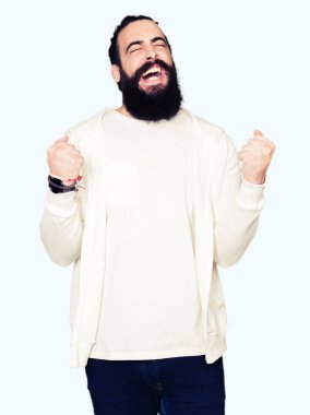 Gülümseyen ve başarı için çığlık genç adamla uzun saç ve sakal sportif sweatshirt çok mutlu ve heyecanlı kaldırdı, silah ile kazanan jest yapıyor giyiyor. Kutlama kavramı.