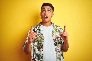 Tatil üzerinde genç Brezilyalı adam yazlık giyen yaz çiçek gömlek üzerinde izole sarı arka plan şaşırdı ve bakıyor ve parmaklar ve yükseltilmiş kollar ile işaret şaşırttı.