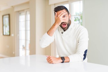 Yakışıklı İspanyol adam evde rahat beyaz kazak giyiyor şok kapsayan yüz ve gözleri el ile, utanmış ifade ile parmaklarla bakıyor.