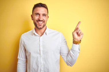 Yüze büyük bir gülümseme ile sarı izole arka plan üzerinde zarif beyaz gömlek giyen genç yakışıklı adam, kamera bakarak yan el ve parmak ile işaret.
