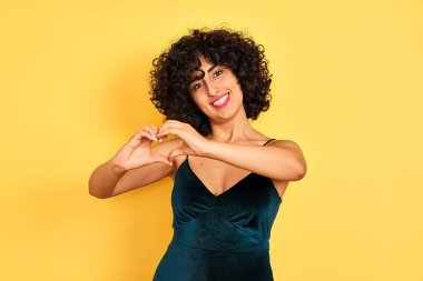Kıvırcık saçlı genç arap kadın izole sarı arka plan üzerinde zarif elbise giyen aşk elleri ile kalp sembolü şekli yapıyor gülümseyerek. Romantik kavram.