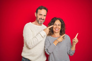 Güzel orta yaş çift izole kırmızı arka plan üzerinde kış kazak giyiyor gülümseyen ve iki el ve parmak tarafına işaret kamera bakıyor.
