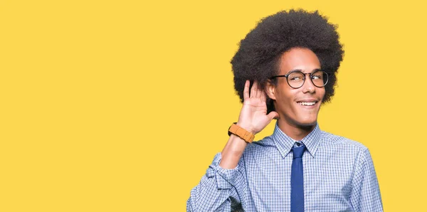 Young african american business man with afro hair wearing glasses smiling with hand over ear listening an hearing to rumor or gossip. Deafness concept.