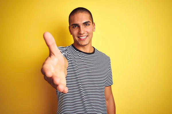 黄色の孤立した背景の上にストライプのTシャツを着た若いハンサムな男は 挨拶と歓迎として握手を提供する友好的な笑顔 ビジネスの成功 — ストック写真