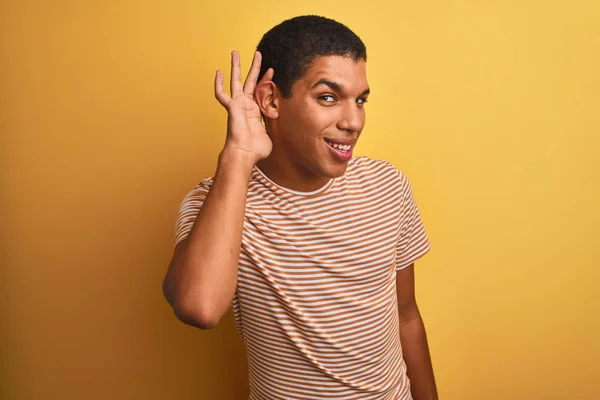 年轻的英俊阿拉伯男子穿着条纹T恤站在孤立的黄色背景微笑 手在耳朵上听听谣言或八卦 耳聋概念 — 图库照片