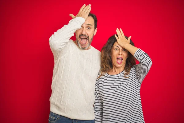 Beautiful middle age couple wearing winter sweater over isolated red background surprised with hand on head for mistake, remember error. Forgot, bad memory concept.