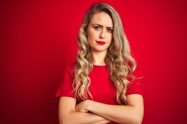 Kırmızı izole arka plan üzerinde ayakta temel t-shirt giyen genç güzel kadın şüpheci ve sinir, kesişmiş kollar ile yüz üzerinde ifade onaylamıyor. Negatif kişi.
