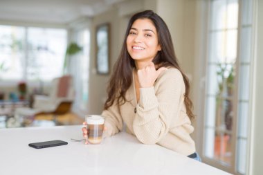 Genç kadın evde bir fincan kahve içme işaret ve mutlu yüz gülümseyen yanında başparmak ile gösteren