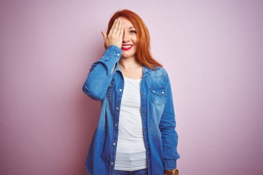 Youg güzel kızıl saçlı kadın kot gömlek ayakta izole pembe arka plan üzerine el ile bir göz kapsayan, yüz ve sürpriz duygu kendine güvenen gülümseme giyen.