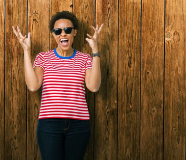 Güzel genç Afro-Amerikan kadın üzerinde güneş gözlüğü takıyor deli kutluyor arka plan izole ve silah ile başarı için yükseltilmiş ve açık gözleri heyecanlı çığlık hayrete düşürdü. Kazanan kavramı