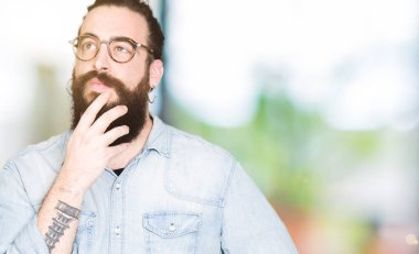 Uzun saç ve sakal gözlük el ile çene düşünme hakkında soru, dalgın ifade üzerine genç hipster adam. Düşünceli yüzüyle gülümsüyor. Şüphe kavramı.