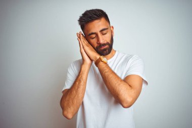 Kapalı gözleri ile gülümseyen iken izole beyaz arka plan uyku yorgun hayal ve eller ile poz üzerinde ayakta t-shirt giyen genç Hintli adam.