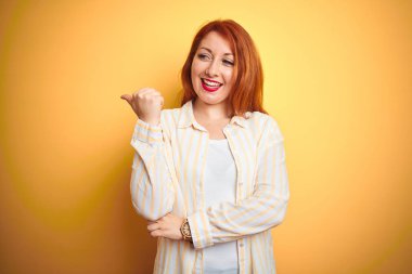 Mutlu yüz görünümlü ve başparmak ile yan işaret ile gülümseyen izole sarı arka plan üzerinde ayakta çizgili gömlek giyen güzel Redhead kadın.
