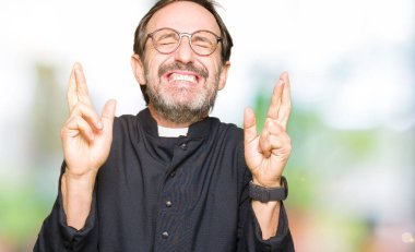 Orta yaş rahip adam geçiş parmakları umut ve gözleri kapalı ile gülümseyen Katolik bornoz giymiş. Şans ve batıl kavramı.