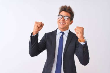 Genç yakışıklı işadamı izole beyaz arka plan üzerinde takım elbise ve gözlük giyen çok mutlu ve heyecanlı kolları kaldırdı ile kazanan jest yapıyor, gülümseyerek ve başarı için çığlık. Kutlama konsepti.