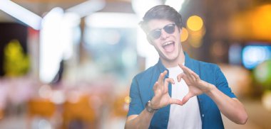 Genç yakışıklı adam aşık kalp simgesi ve elleriyle şekil gösteren gülümseyen izole arka plan üzerinde güneş gözlüğü takıyor. Romantik kavramı.