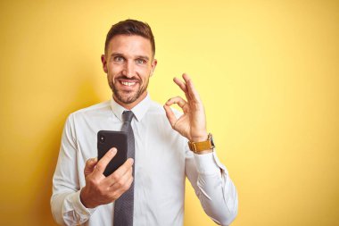 Sarı izole arka plan üzerinden akıllı telefon kullanarak genç yakışıklı iş adamı parmaklar ile ok işareti yapıyor, mükemmel sembolü