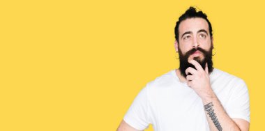 Uzun saç ve sakal çene düşünme hakkında soru, dalgın ifade üzerine el ile rahat beyaz t-shirt giyen genç hipster adam. Düşünceli yüzüyle gülümsüyor. Şüphe kavramı.